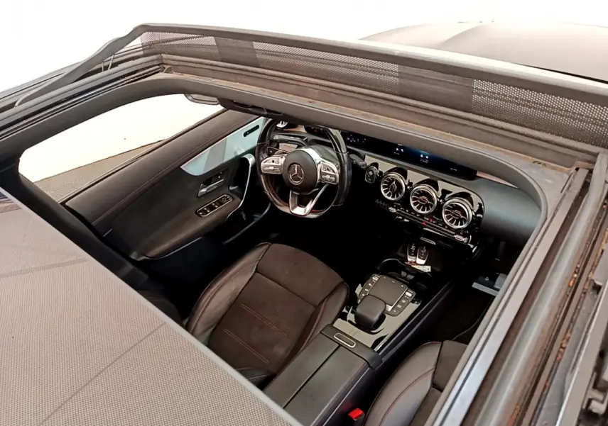 Vue intérieure par le toit ouvrant du poste de conduite noir avec volant multifonction et aérateurs ronds du Mercedes CLA 200 d AMG Line.