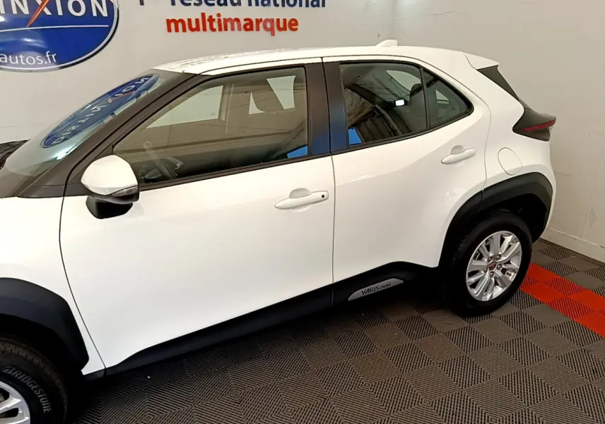 Profil côté gauche d'une Toyota Yaris Cross blanche 2022 avec logo sur bas de caisse, en intérieur showroom.