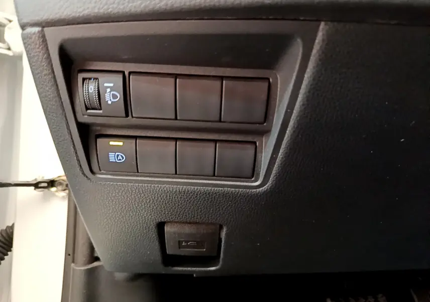 Gros plan sur les commandes d'éclairage et un port USB sous le tableau de bord côté conducteur d'une Toyota Yaris Cross blanche.
