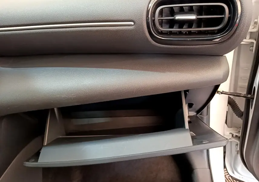 Gros plan sur la boîte à gants ouverte côté passager du Toyota Yaris Cross blanc, montrant l'intérieur noir du tableau de bord.