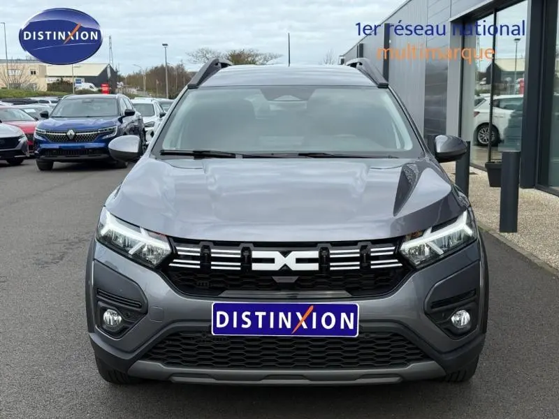 Vue avant d'un Dacia Jogger gris 2025 avec calandre noire et logo distinctif, stationné devant un showroom.