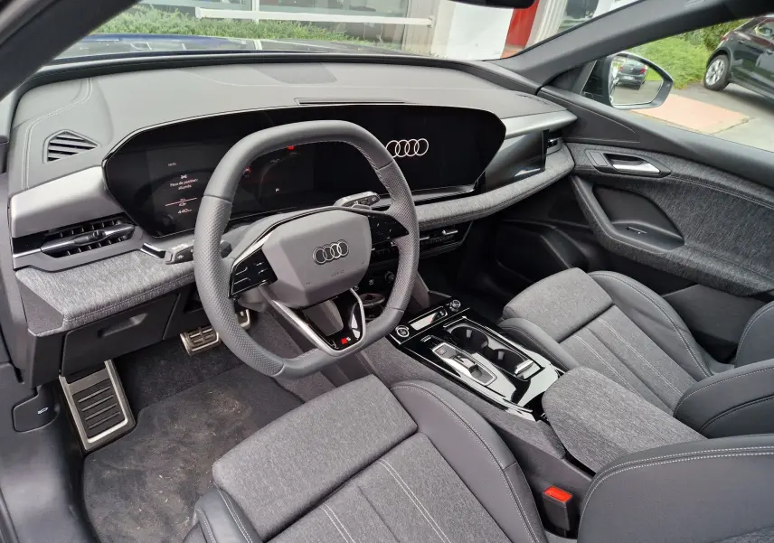 Intérieur de l'Audi Q6 e-tron Performance S-Line 2025, vue côté conducteur, tableau de bord digital et sièges gris tissu et cuir.