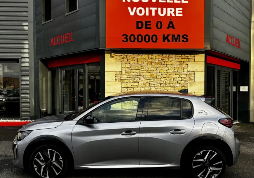 Profil droit de la Peugeot 208 Hybrid 110 e-DCS6 GT gris clair, avec jantes noires et toit noir, devant un bâtiment commercial.