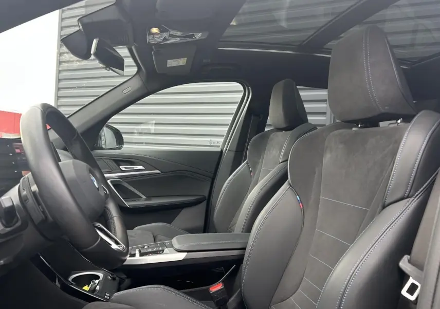 Intérieur BMW X1 2025 en vue côté gauche, sièges sport en alcantara noir avec surpiqûres bleues et toit panoramique.