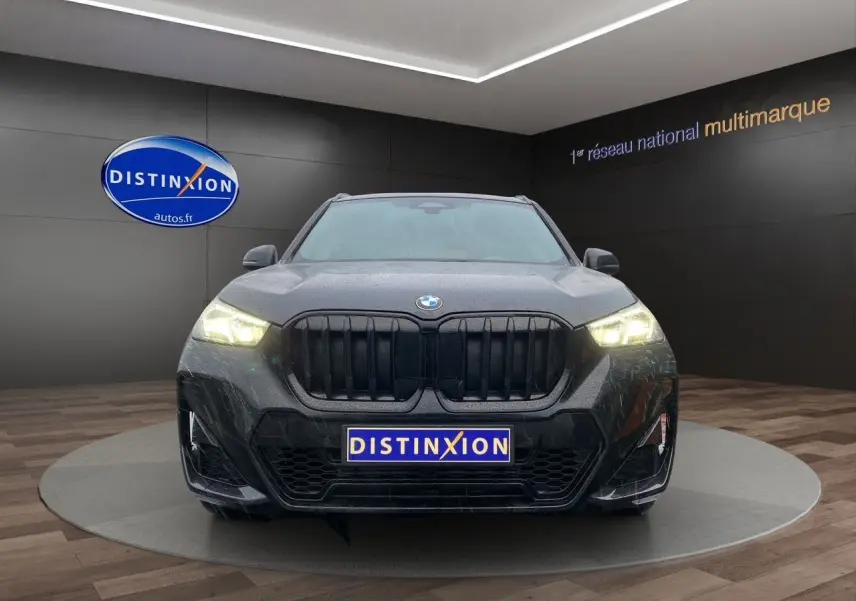 Vue frontale d'un BMW X1 XDRIVE 20D M SPORT noir Saphir avec phares allumés dans un showroom.