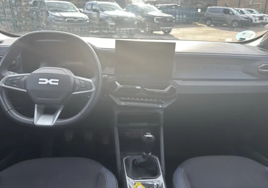 Vue intérieure frontale du tableau de bord du Dacia Duster 2025 avec volant cuir, écran tactile et levier de vitesses manuel.