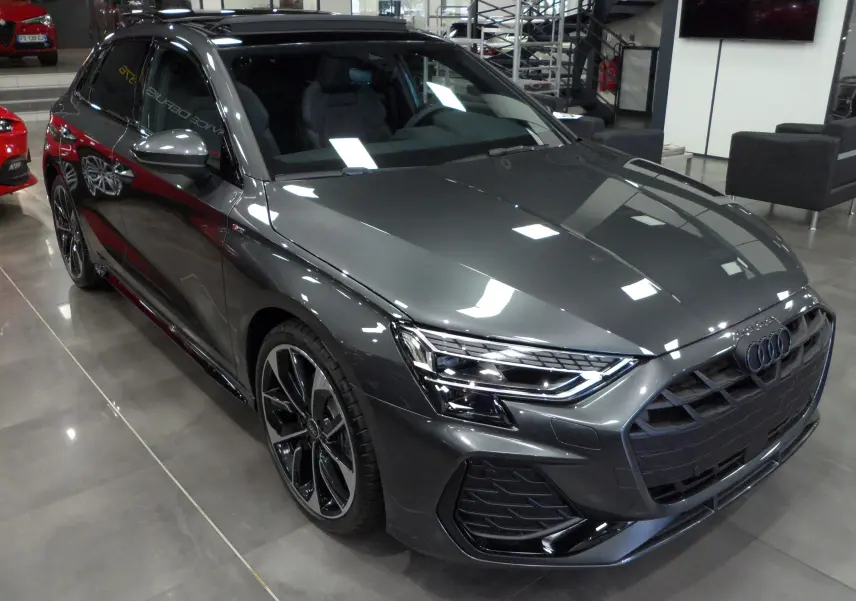 Audi A3 Sportback gris Daytona vue 3/4 avant droit en showroom, avec jantes noires bi-ton et calandre S-Line.