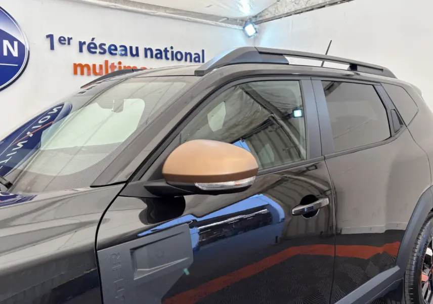 Vue latérale gauche du Dacia Duster noir avec rétroviseur couleur bronze et barres de toit noires visibles.