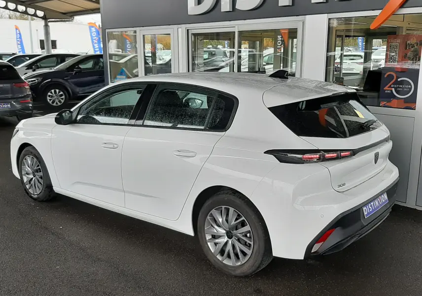 Vue 3/4 arrière droite d'une Peugeot 308 blanche banquise garée devant un showroom avec feux arrière LED distinctifs.