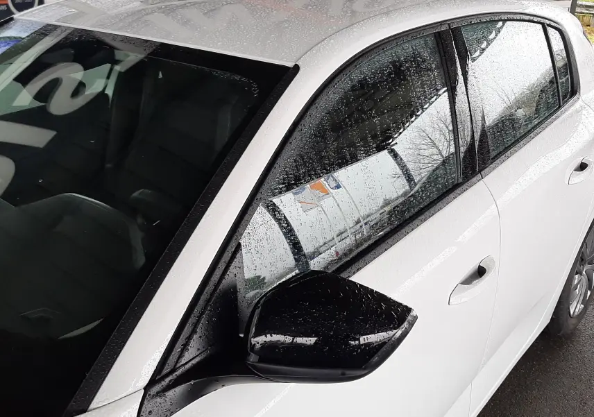 Vue côté gauche avant d'une Peugeot 308 blanche banquise avec rétroviseur noir sous la pluie, détails de la porte et vitre visibles.