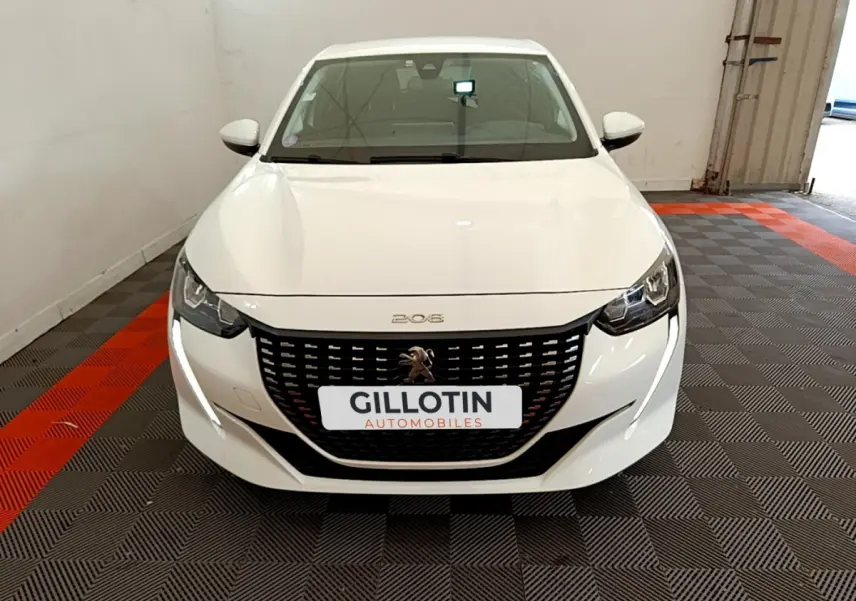 Vue frontale d'une Peugeot 208 blanche avec calandre noire et feux LED, stationnée sur un sol à damiers gris et rouge.