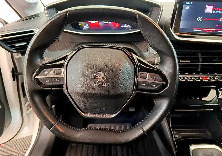 Vue rapprochée du volant cuir noir de la Peugeot 208 blanche, avec commandes intégrées et tableau de bord numérique visible.