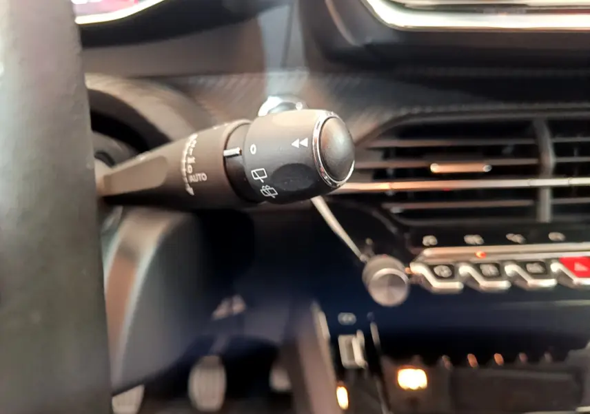 Gros plan sur la commande d'essuie-glace à gauche du volant dans l'habitacle sombre d'une Peugeot 208 blanche.