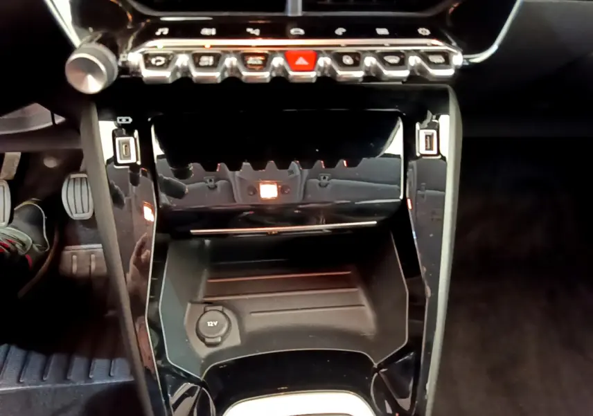Vue rapprochée de la console centrale noire du Peugeot 208 blanc, avec prise 12V et commandes tactiles au-dessus.