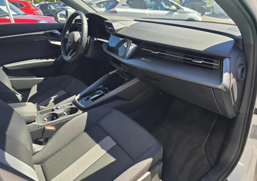 Vue intérieure côté passager d'une Audi A3 Sportback 2024 blanche, avec tableau de bord noir et sièges tissu gris foncé.