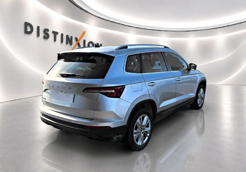Vue 3/4 arrière droite d’un Skoda Karoq gris 2025 avec barres de toit et feux arrière LED dans un showroom lumineux.