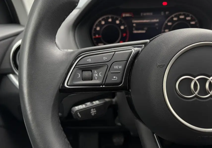 Gros plan sur le volant cuir multifonction de l'Audi Q2 30 TFSI S-Line avec commandes et tableau de bord numérique flou.