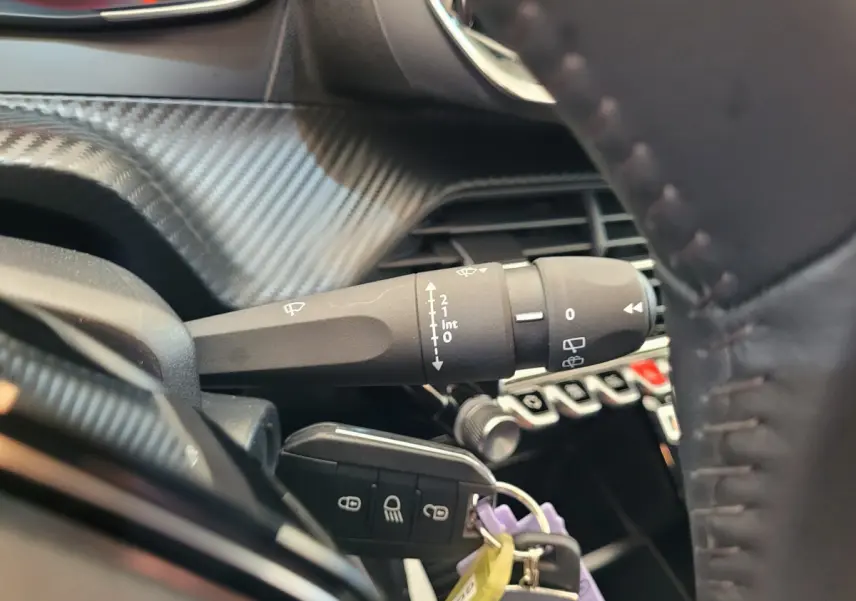 Gros plan sur la manette d'essuie-glace et clé de contact dans l'habitacle d'une Peugeot 208 gris foncé, volant cuir visible.
