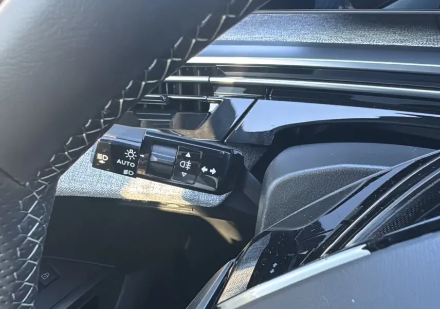Détail du commodo d’éclairage noir brillant côté gauche du volant dans l’habitacle du Peugeot 3008 New Hybrid 2025.
