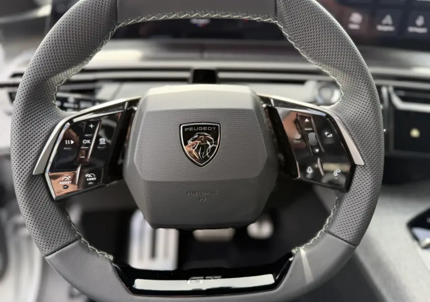 Volant en cuir perforé gris avec commandes intégrées et logo Peugeot au centre, vue frontale rapprochée.