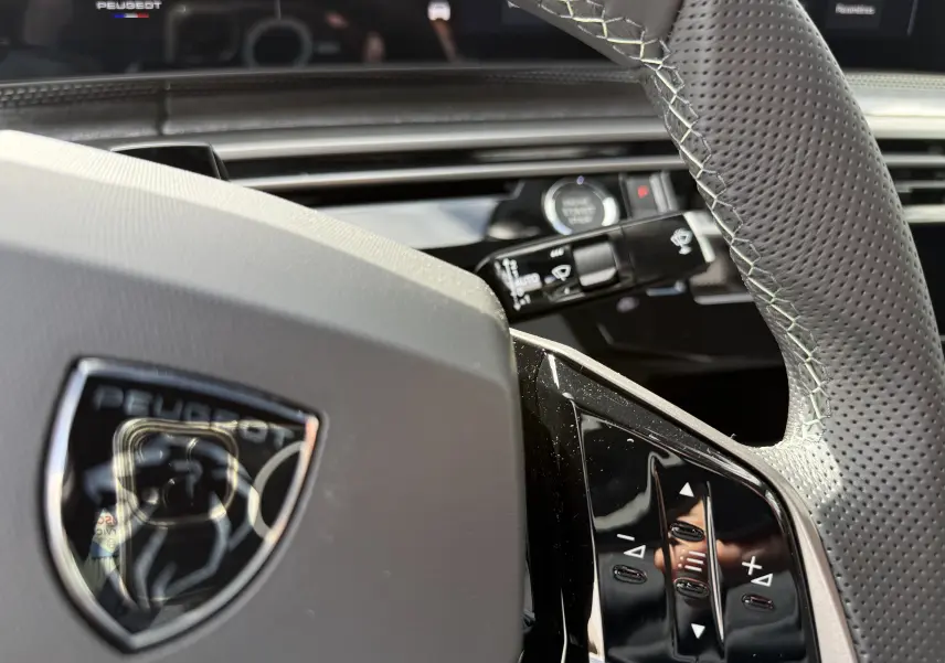 Gros plan sur le volant en cuir perforé gris avec logo Peugeot, commandes intégrées et tableau de bord numérique en arrière-plan.