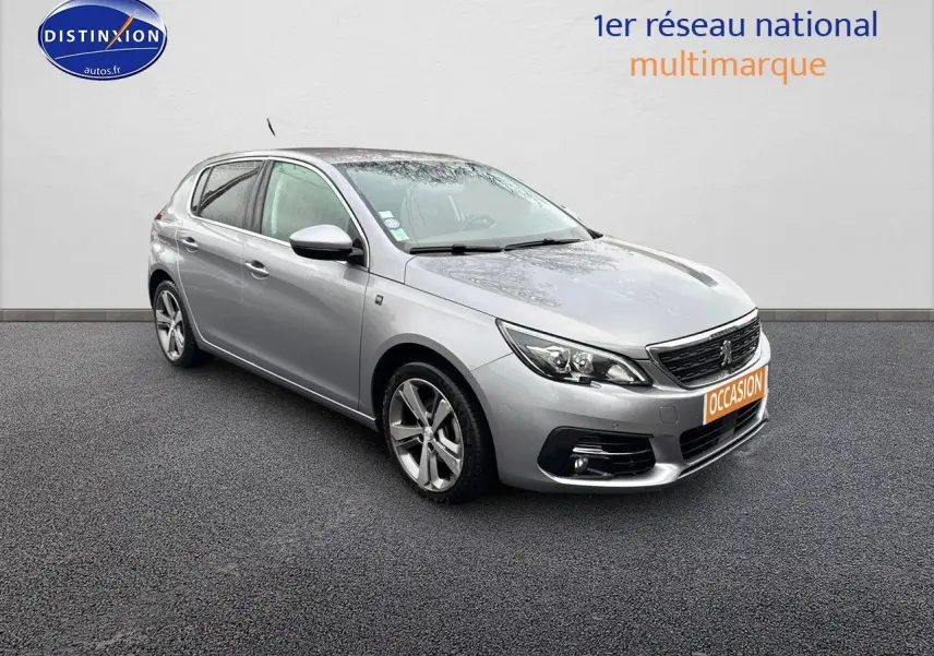 Peugeot 308 gris Artense vue 3/4 avant droit sur sol gris avec plaque orange "OCCASION" visible.