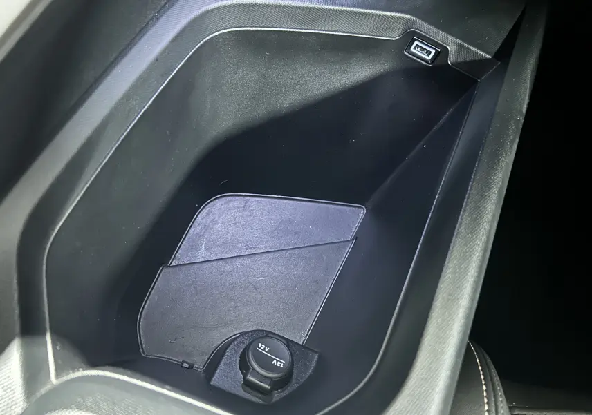 Gros plan sur la console centrale noire du Peugeot 5008 hybride 2025, montrant une prise 12V et un port USB.