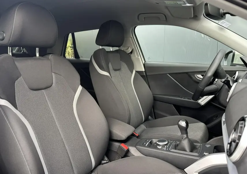 Vue intérieure côté conducteur et passager avant de l'Audi Q2 30 TFSI S-Line 2024 avec sièges sport tissu gris et levier de vitesses manuel.