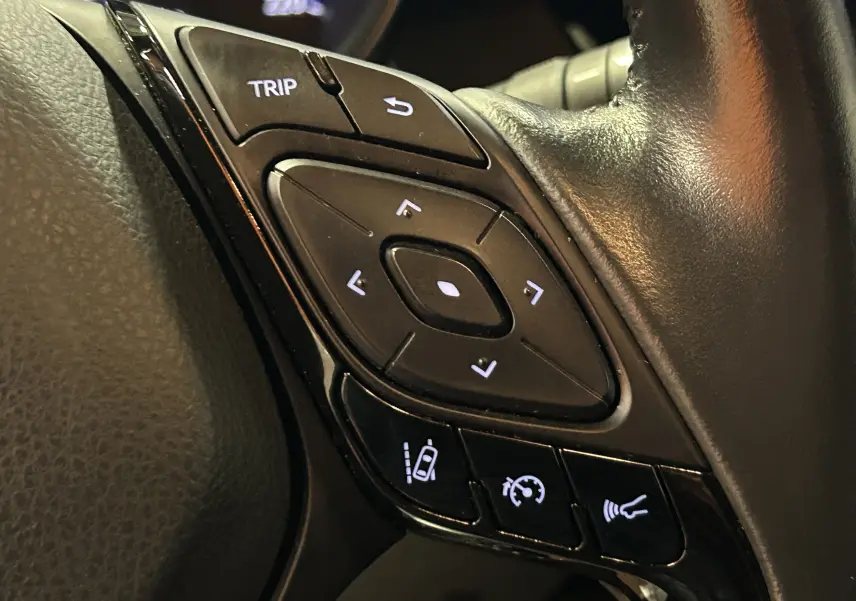 Gros plan sur les commandes du volant en cuir noir du Toyota C-HR Hybride 122h Edition 2WD, boutons rétroéclairés blancs.