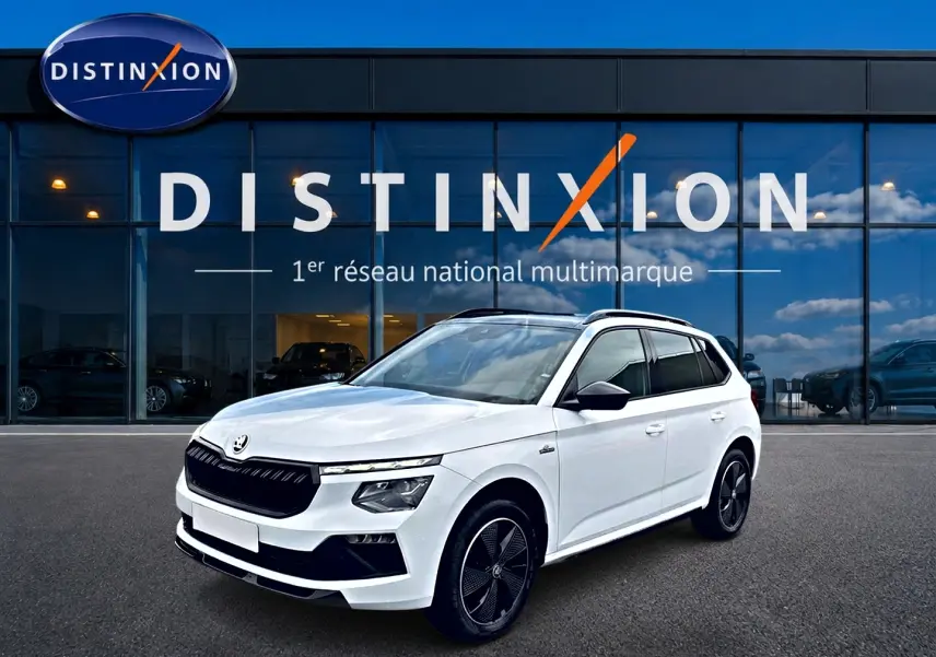 Vue 3/4 avant d'un Skoda Kamiq blanc nacré avec jantes noires et toit panoramique devant une vitrine Distinxion.