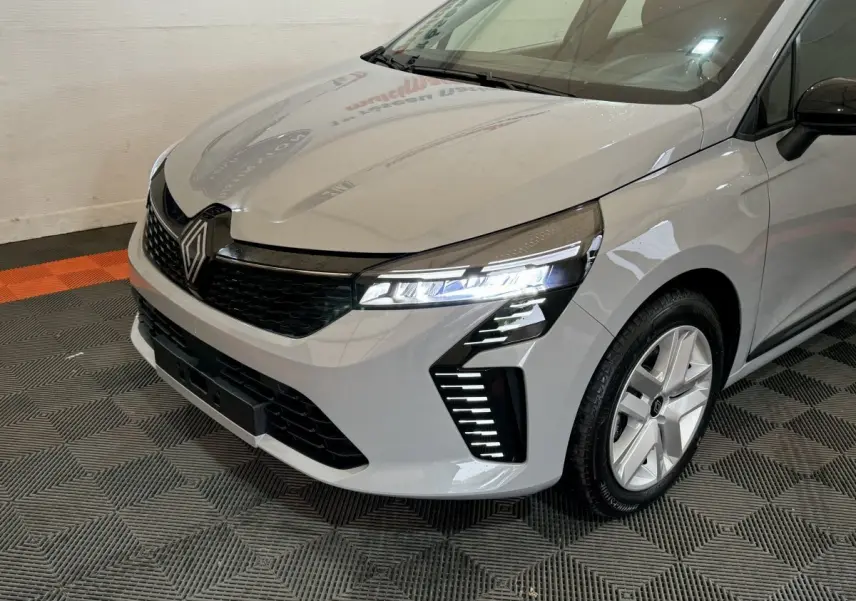 Vue 3/4 avant droit d'une Renault Clio gris clair 2025 avec phares LED allumés et calandre noire.