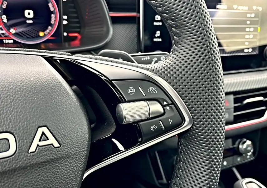 Gros plan sur le volant perforé noir avec commandes multifonctions du Skoda Kamiq blanc nacré, tableau de bord numérique en arrière-plan.