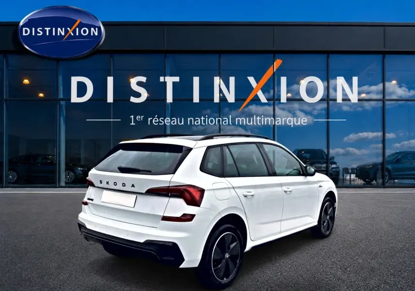 Vue 3/4 arrière droite d'un Skoda Kamiq blanc nacré avec jantes noires et toit panoramique, devant un showroom Distinxion.