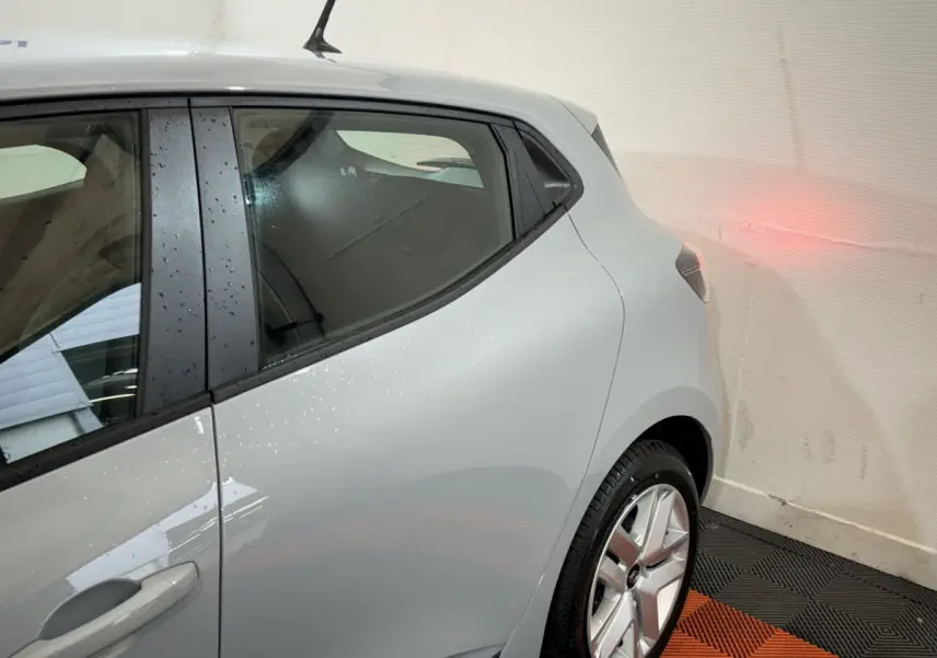 Vue côté gauche arrière d'une Renault Clio gris clair avec jante alliage et portière arrière visible en intérieur fermé.