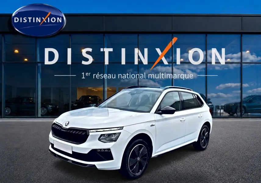 Vue 3/4 avant droite d'un Skoda Kamiq blanc nacré avec jantes noires et toit panoramique devant un showroom Distinxion.