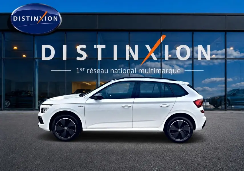 Profil droit d'un Skoda Kamiq blanc nacré 2026 avec jantes noires et toit panoramique visible, devant une vitrine Distinxion.