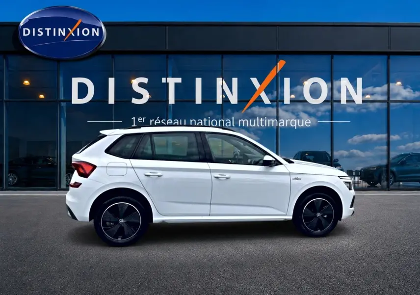 Vue latérale droite d'un Skoda Kamiq blanc nacré avec jantes noires devant un bâtiment vitré Distinxion.