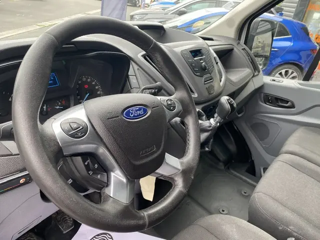 Vue intérieure côté conducteur du Ford Transit Fourgon blanc 2019, volant multifonctions et tableau de bord avec commandes intégrées.