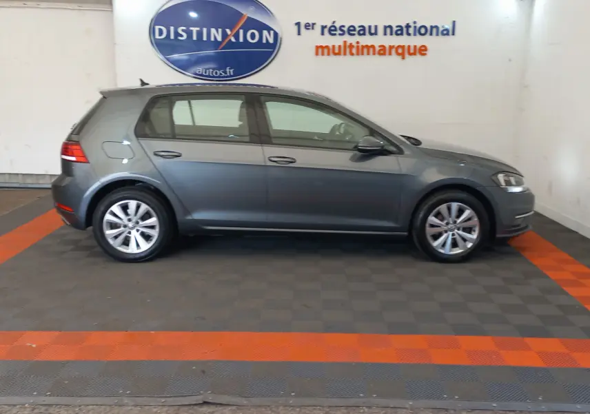 Profil côté droit d'une Volkswagen Golf gris foncé 2019 garée en intérieur sur sol à damiers avec logo Distinxion en arrière-plan.