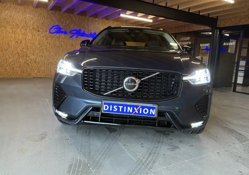 Vue frontale d’un Volvo XC60 bleu avec calandre noire laquée et phares allumés en intérieur showroom.