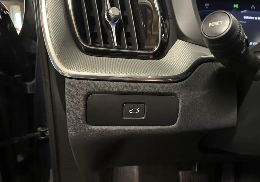Gros plan sur le bouton d'ouverture du hayon dans l'habitacle noir du Volvo XC60 2023 côté conducteur.
