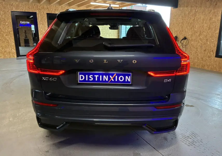 Vue arrière d’un Volvo XC60 bleu foncé 2023 avec feux LED allumés et logo distinctif sur un mur bois.