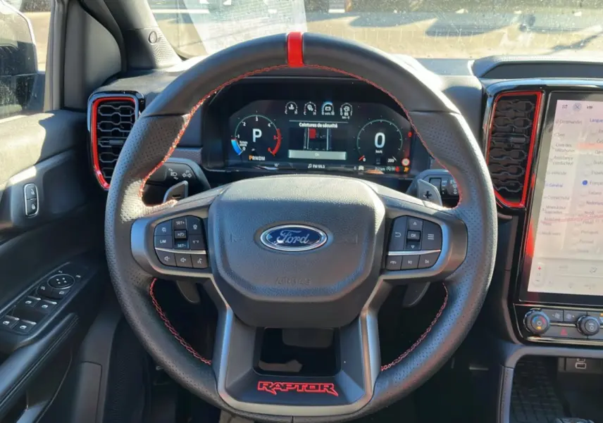Vue intérieure centrée sur le volant cuir noir avec surpiqûres rouges et tableau de bord numérique du Ford Ranger Raptor gris commandeur.