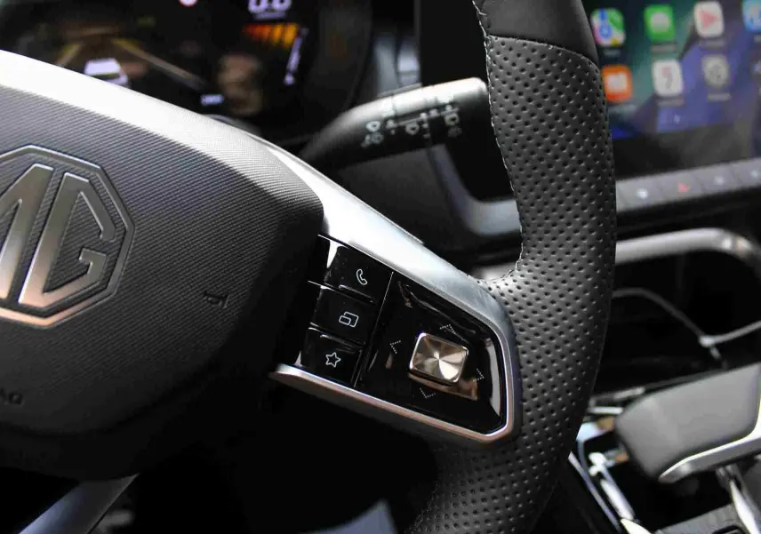 Gros plan sur le volant perforé et les commandes multifonctions du MG ZS hybride, intérieur gris et noir visible.