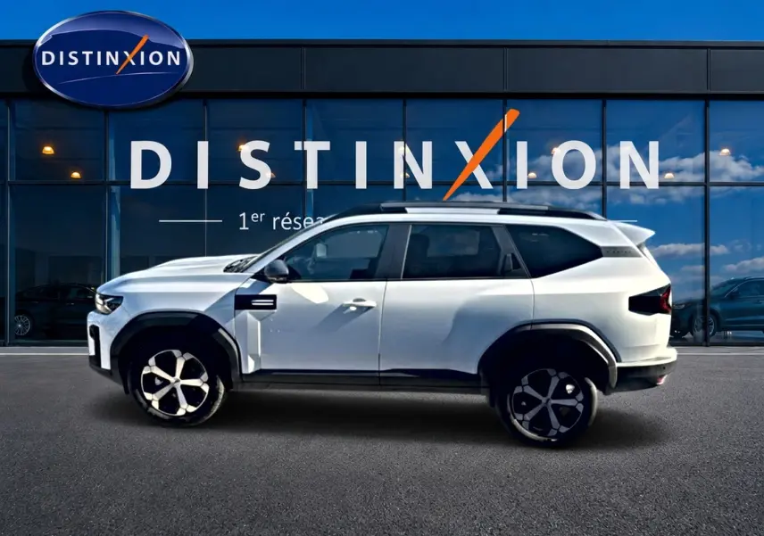 Vue de profil côté gauche d’un Dacia Bigster blanc 2026 avec barres de toit et jantes alu devant une vitrine Distinxion.