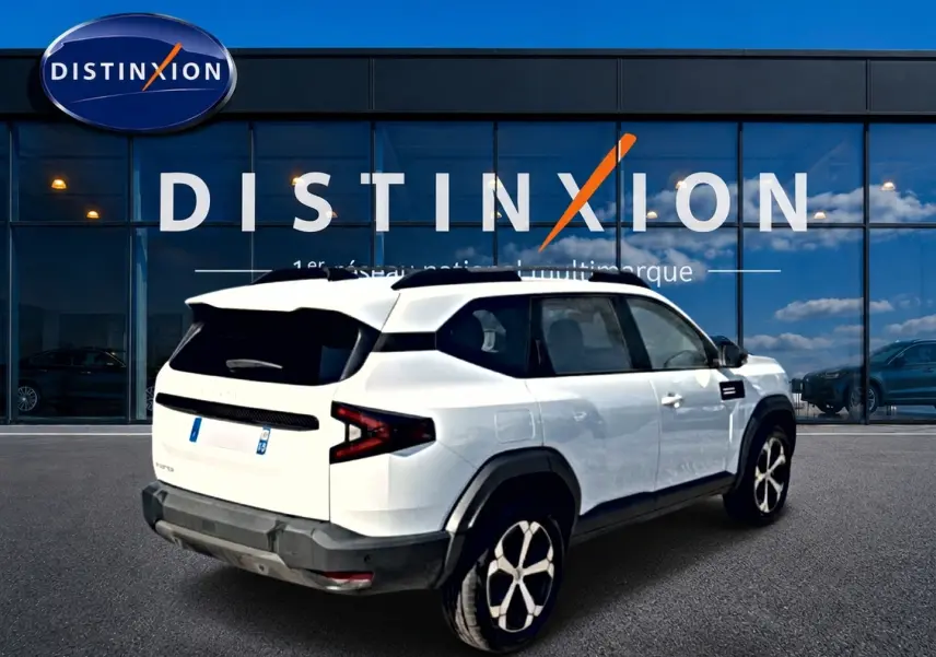 Vue 3/4 arrière droite du SUV blanc Dacia Bigster 2026 avec jantes alu et toit noir devant un showroom Distinxion.