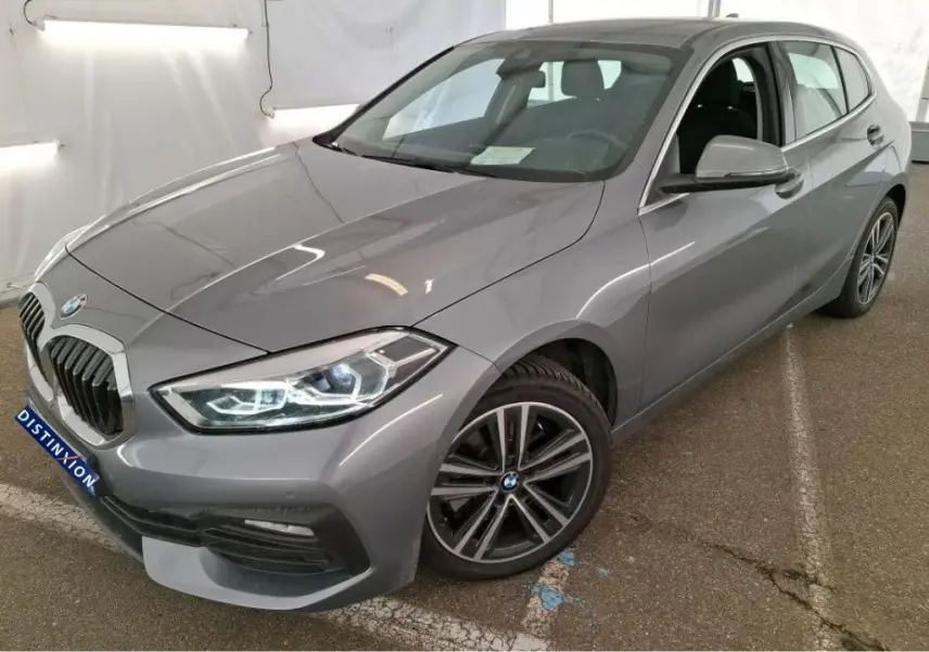 BMW Série 1 gris vue 3/4 avant droit, phares allumés et jantes alliage distinctives, stationnée en intérieur.