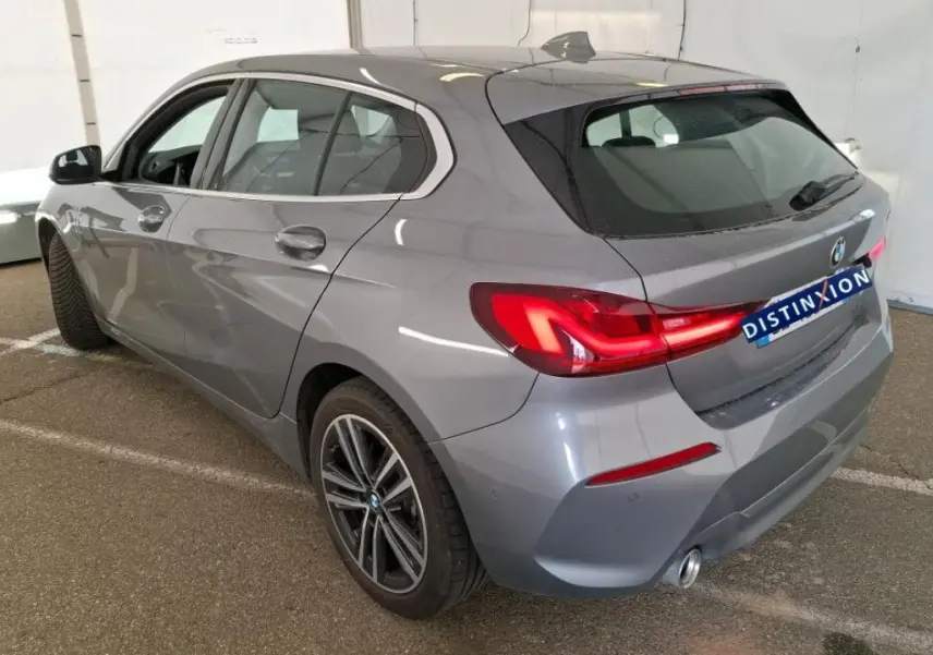 BMW Série 1 116d Business Design gris vue 3/4 arrière droit, feux arrière allumés et jantes alliage visibles.