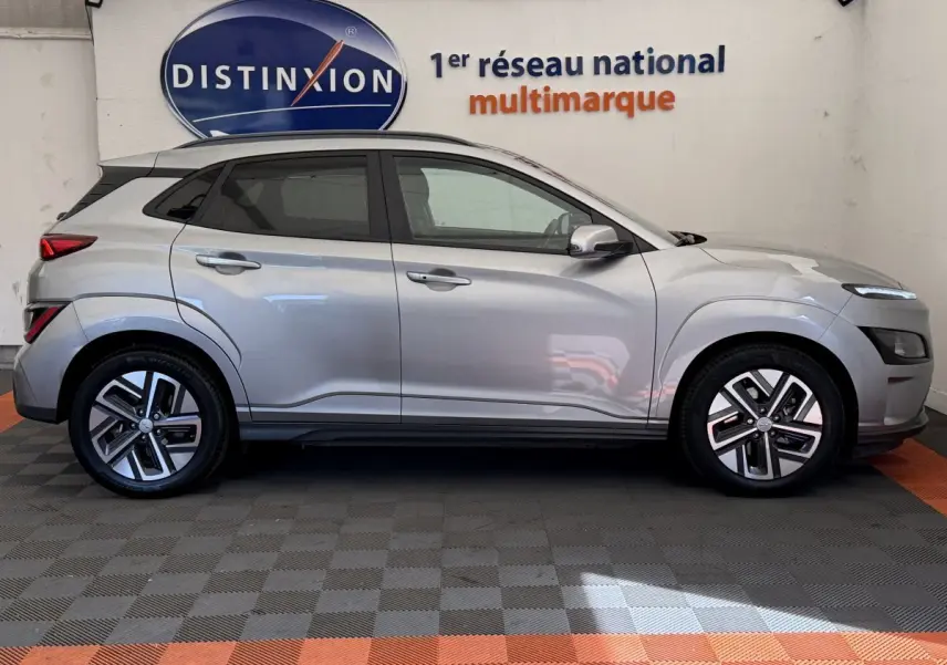 Vue de profil côté gauche d'un Hyundai Kona électrique gris clair garé en intérieur avec logo Distinxion en arrière-plan.