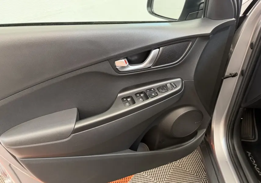 Vue intérieure côté gauche de la porte avant gris clair du Hyundai Kona électrique avec commandes de vitres et haut-parleur intégré.
